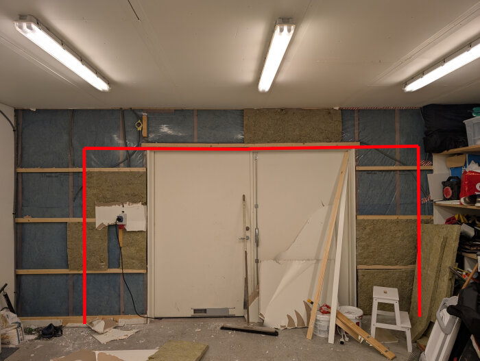 Interiör av garage med en vit slagport, gipsskivor och isolering borttagna. Röda linjer markerar den planerade öppningens bredd för ny garageport.