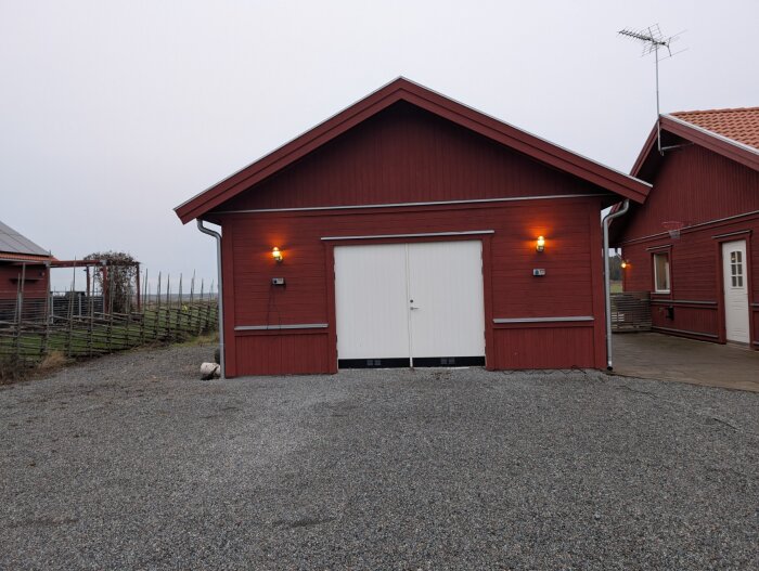 Rött garage med vit slagport på gårdsplan, belysning ovanför dörrarna. Planerar att ersätta porten med en bredare takskjutsport.