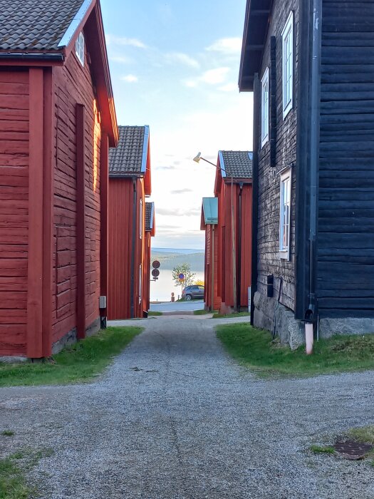 Grusgång mellan röda och svarta trähus med havsutsikt i bakgrunden och en parkerad bil nära vattnet under blå himmel.
