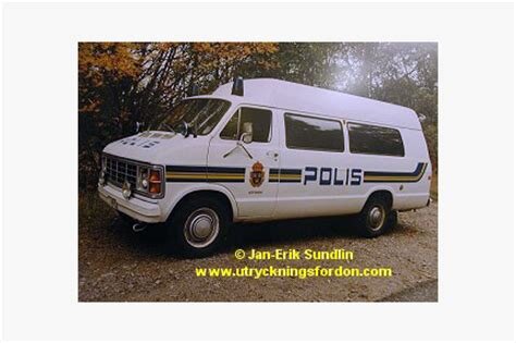 En äldre vit polisbil med texten "Polis" på sidan, parkerad på en grusväg omgiven av träd.