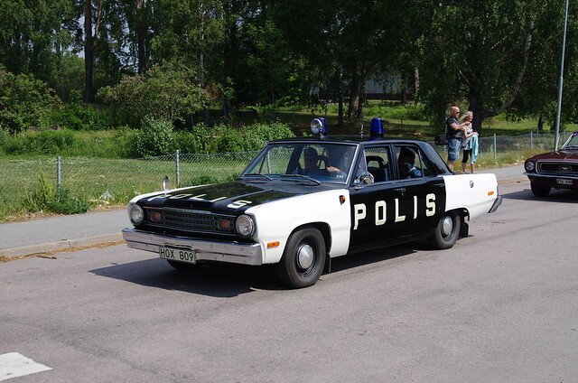 En äldre polisbilsmodell, Dodge, med texten "POLIS" på sidan, körs i dagsljus på en landsväg med grönska i bakgrunden.