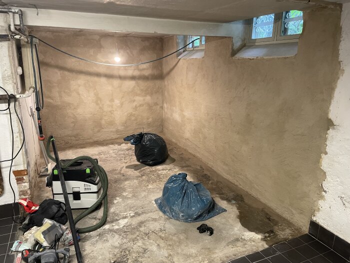 Rum med grovputsade väggar, verktyg och plastpåsar på golvet samt en taklampa och fönster som ger ljusinsläpp.