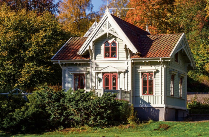 Ett äldre hus med grön fasad och rödbruna fönsterluckor, omgivet av frodig vegetation och höstfärger i bakgrunden.