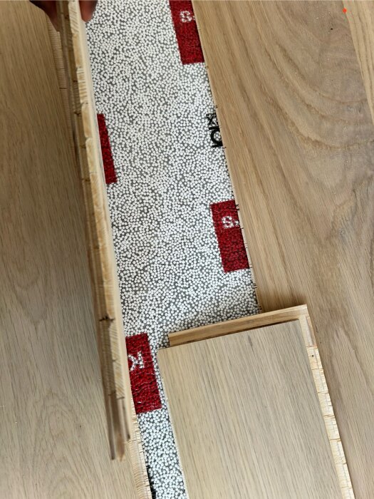 Parkettgolvbräda lyfts för att visa vad som finns under den, inklusive ljudisolerande material med vita och röda prickar.