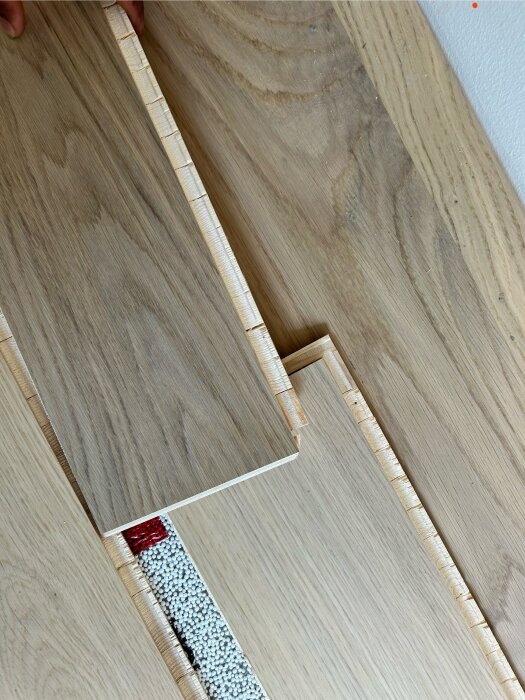 Parkettgolv med nya brädor som ska ersätta de skadade. Under parkettgolvet syns ett lager av isoleringsmaterial och ett hål där golvbrädor saknas.