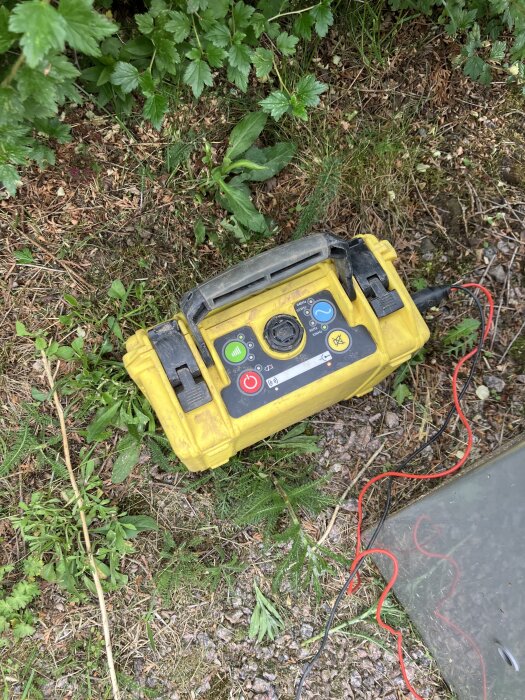 Gul mätlåda med sladdar placerad på marken bredvid växtlighet, intill ett öppnat elskåp.