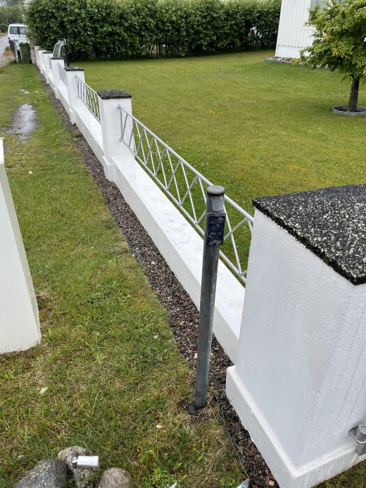 En vitmålad markmur av betong med metallstaket, stående mellan staketstolpar av puts, bredvid en gräsmatta. En grå stolpe står framför muren.