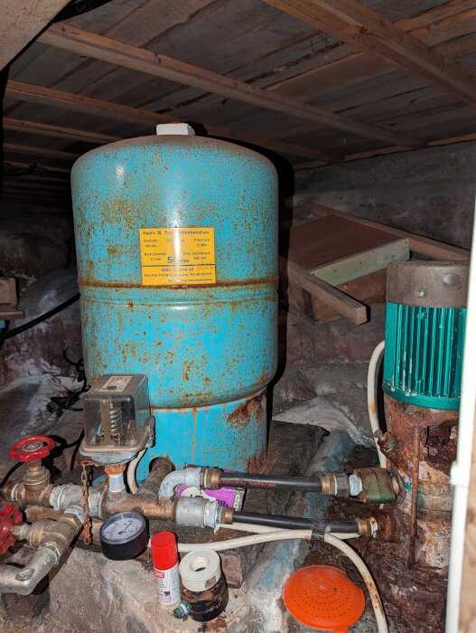 Rostig hydrofor och pumpinstallation i källare, med ventiler, tryckmätare och kopplingar på en klarröd varmvattenledning. Ventil med kedja.