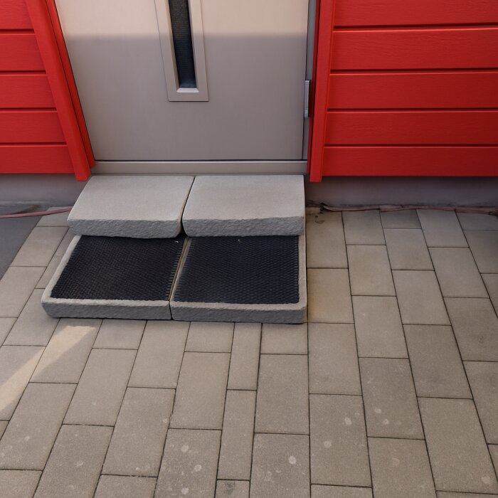 Grå betongplattor placerade vid en röd och beige entré, med två svarta gummimattor mellan plattorna och dörren.