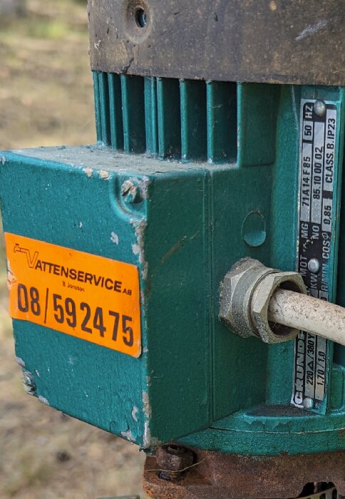 Närbild av en gammal, sliten pump med grönt hölje och en orange etikett från ett serviceföretag, monterad med rördelar.