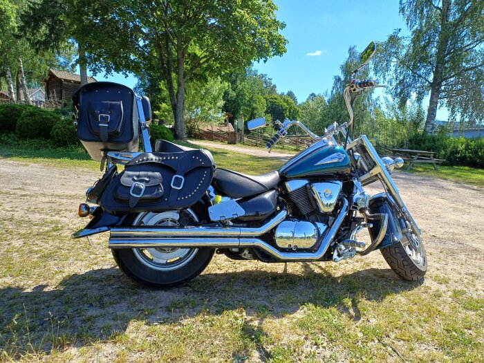 En cruiser-motorcykel med sadelväskor, parkerad på grus med grönska i bakgrunden.