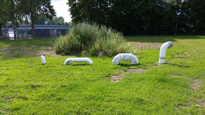 Ventilationsrör arrangerade i gräs på en park eller trädgård, med buskar och ett staket i bakgrunden.