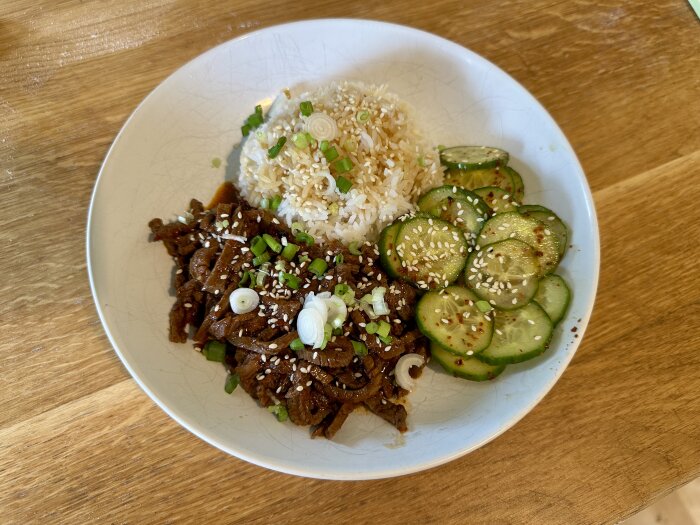 Strimlad lövbiff i sötstark sås med kokt ris och gurkskivor, serverat på tallrik, garnerat med sesamfrön och salladslök.