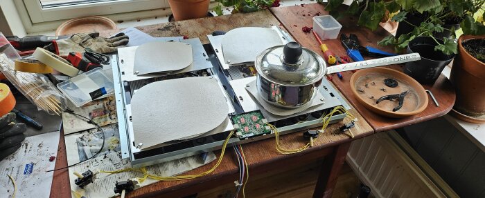 Nedmonterad spis på ett bord med kastrull ovanpå, omgiven av verktyg och sladdar, samt växter i krukor i bakgrunden.