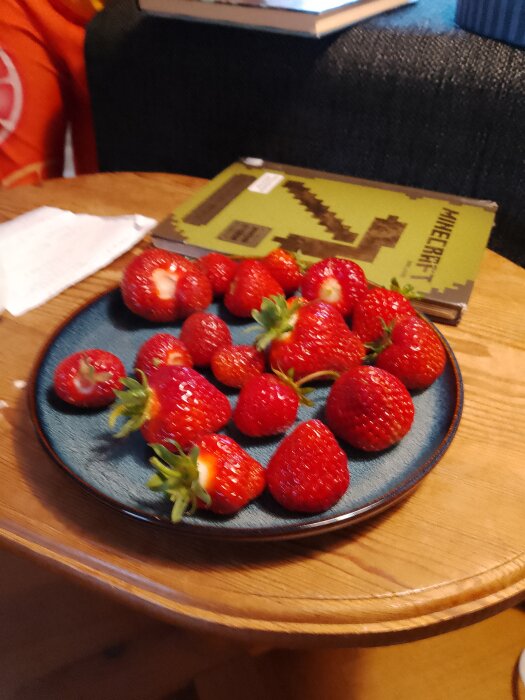 En suddig bild av en tallrik med nyplockade jordgubbar på ett bord, bredvid en bok.