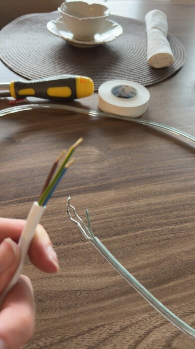 Närbild av en hand som håller en kabel med färgade ledningar jämte en genomskinlig kabel på ett bord med verktyg i bakgrunden.