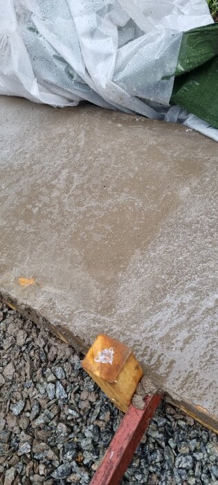 Betongsula under härdning, täckt delvis av plast för skydd. Grus syns vid sidan och en trästöd fäster plasten vid ena hörnet.