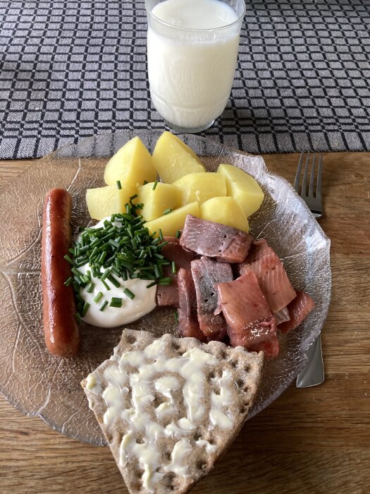Matjestallrik med potatis, korv, gräddfil toppad med gräslök, sill, och knäckebröd med smör, serverad med ett glas mjölk.
