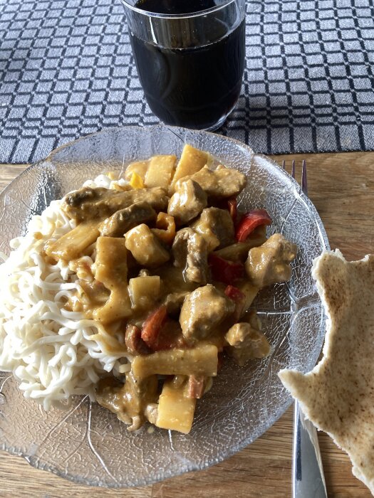 Indisk meat korma med nudlar på en tallrik, serverad med ett glas mörk dryck och en bit bröd.
