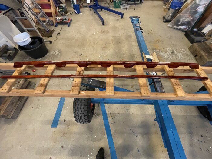 Lackerat grabbräcke i mahogny på en arbetsbänk, del av renovering av träbåt, i ett verkstadsutrymme med verktyg och utrustning omkring.