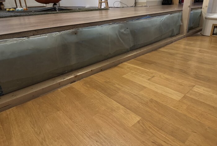 Närbild av en nivåskillnad mellan två golv, med plastskydd och frigolit under laminatgolvet.