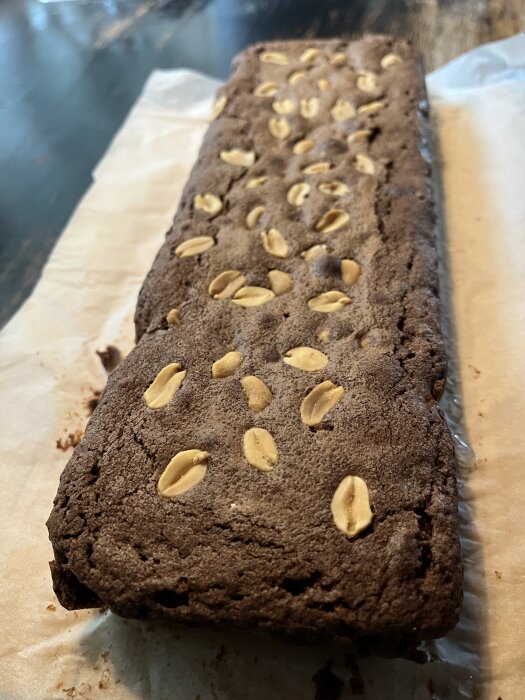 Rektangulär chokladkaka med jordnötter ovanpå, placerad på bakplåtspapper på ett bord.