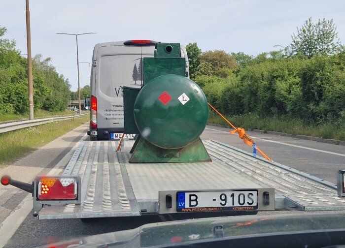 Släpvagn transporterar grön tank fastsatt med orange spännband, bakom vit skåpbil på väg nära grönska.