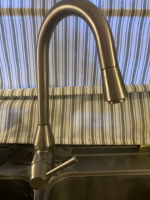 Äldre köksblandare i rostfritt stål, belägen ovanför en diskbänk med randigt draperi i bakgrunden.