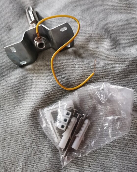 Taklampa fäste med gul kabel och plastpåse innehållande skruvar och pluggar ligger på grått tyg.