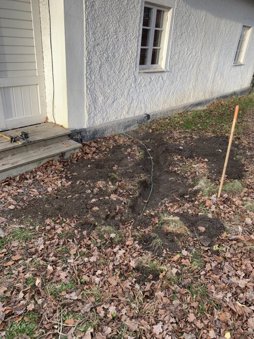 En grävd kanal nära en husvägg med en utstickande grön kabel och en pinne som markerar telefonledningen bland höstlöv.