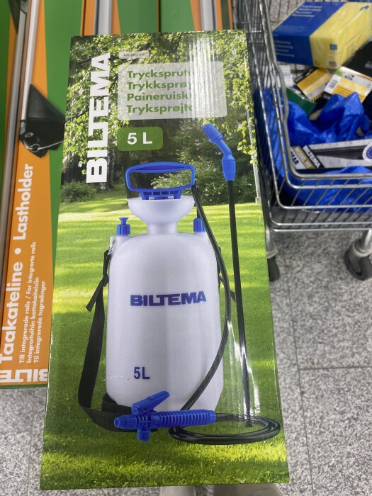 Bild av en tryckspruta från Biltema, kapacitet 5 liter, med blå detaljer och justerbart munstycke. Står i butiksmiljö bredvid en kundkorg.