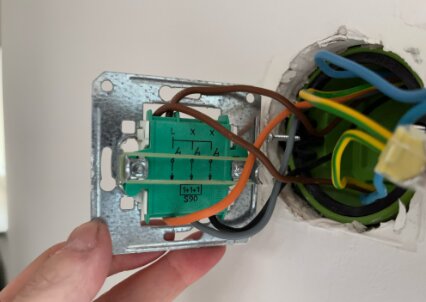 Infälld apparatdosa i vägg med koppla ledningar, hålls av en hand; syns detalj av installation och kabelanslutningar.