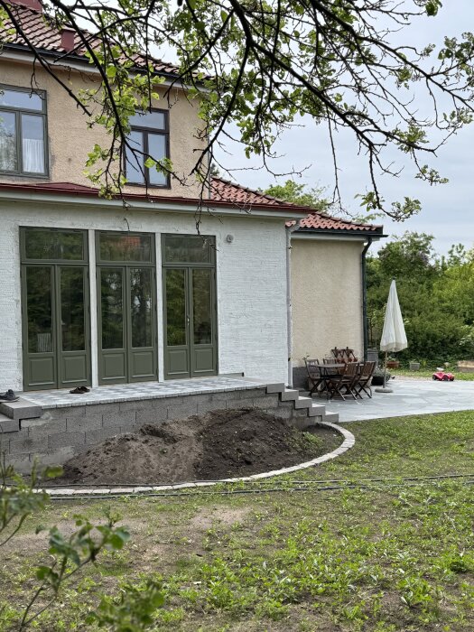 Stenbelagd utbyggnad med trappor på båda sidor; terrass med trädgårdsmöbler och parasoll, omgivna av gräsmatta och buskar.