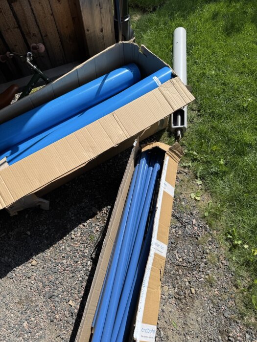 Kartonger med blå borrör och en grå borrdel ligger på gräsmatta vid grusgång.