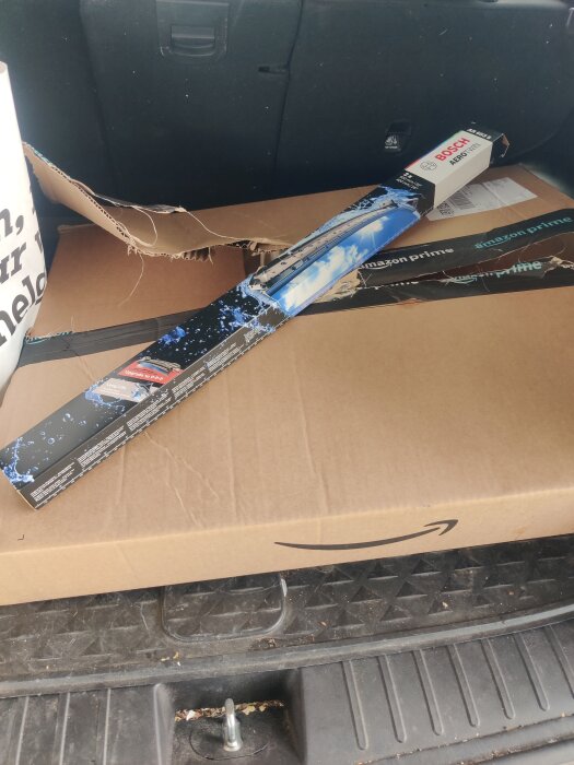 Kartong med stort Amazon-paket i bilens bagageutrymme. Ett paket torkarblad ligger ovanpå, vilket visar ineffektiv förpackning.