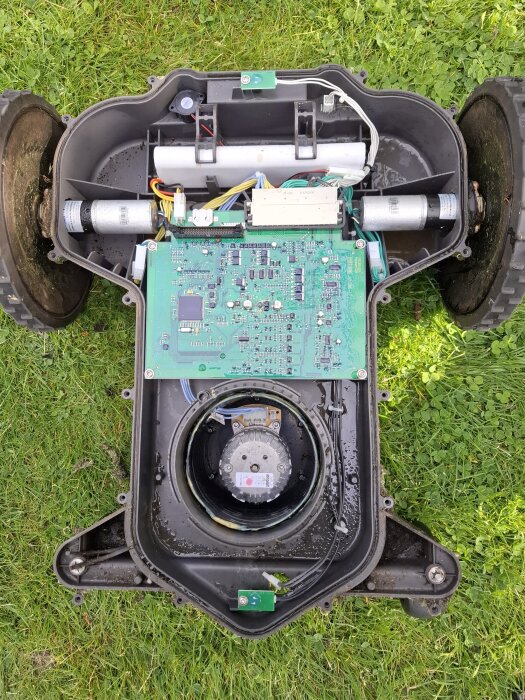 Öppnad robotgräsklippare med synligt moderkort och elektronik, exponerad på en gräsmatta, tecken på fukt inuti höljet.