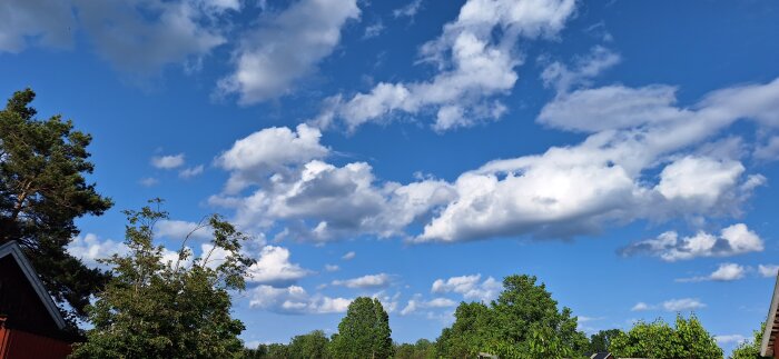 Blå himmel med vita moln och gröna träd i förgrunden, visas ovanför ett hus med rött tak.