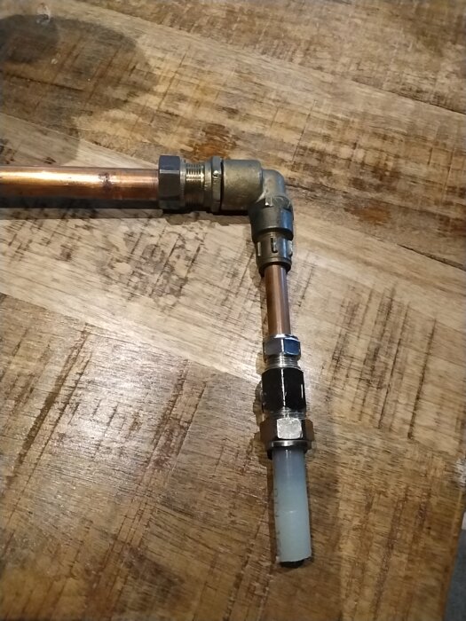 Kopparkoppling med 90-graders vinkel, ansluten till en pex och kopparrör, på ett träbord. Ursprunglig installation av utkastaren för vattenpost.