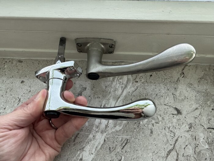 Kromat EFO-61 fönsterhandtag i en hand, bredvid ett äldre monterat handtag, med olika längd på staven som går in i fönstret.