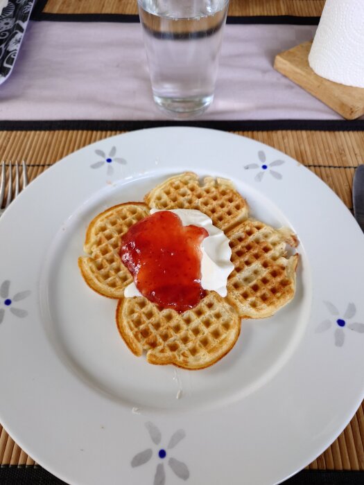 Våffla på tallrik med sylt och grädde, serverad med ett glas vatten och bestick på bord med duk och hushållspappershållare i bakgrunden.