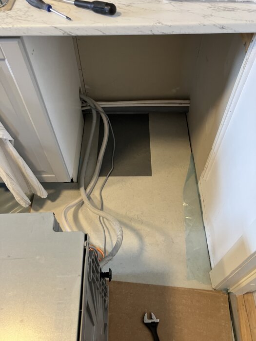 Diskmaskin utdragen under köksbänk med verktyg och slangar, nytt golv delvis lagt, utrymme kvar att täcka.