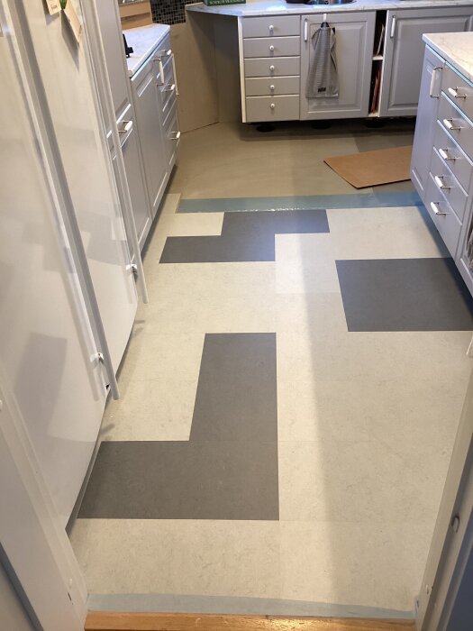 Kök med ljusa skåp, halvlagt golv i olika rektangulära plattor, delvis täckt område, oslipat under diskmaskin.