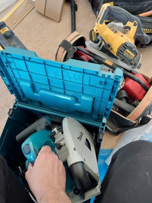 Verktygslåda med handhållen Makita-kapsåg bredvid gul DeWalt-verktyg och byggutrustning på golvet.