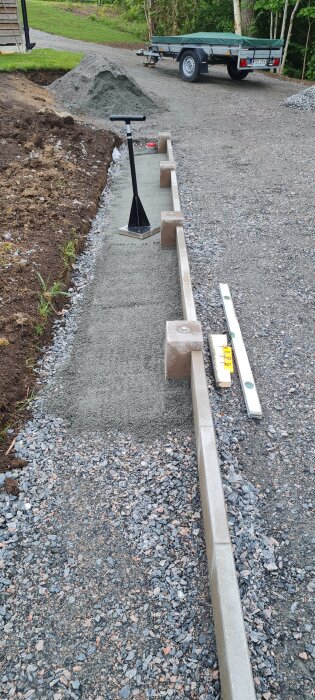 Gångväg med nylagd cement bredvid ett garage, med en kratta och ett vattenpass på sidan, samt grus och en släpkärra i bakgrunden.