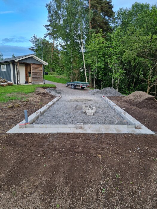 Uppfart med nylagda plattor kring garage, markanläggning pågår, släpvagn i bakgrunden och grönskande skog runtom.