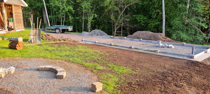 Grus och plattor runt ett garage, med en gräsmatta under anläggning och byggmaterial i en trädgårdsmiljö.
