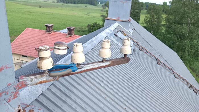 Fyra vita isolatorer fästa på en metallstång på taket, med en rödfärgad byggnad och grönskande åkrar i bakgrunden.