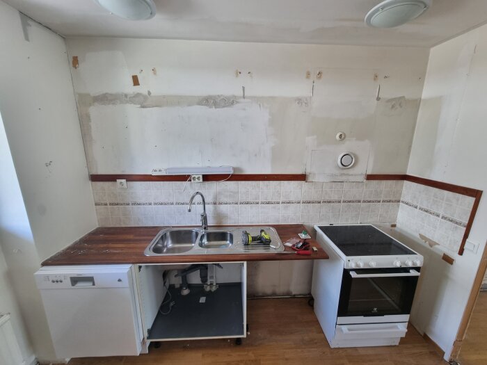 Kök under renovering med synlig diskmaskin, diskho, spis och fläktuttag ovanför arbetsbänk mot kaklad vägg.