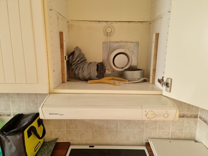 Inuti köksskåpet ovanför spisen visas en borttagen fläktinstallation, med ett flexibelt ventileringsrör och elektriska kablar.