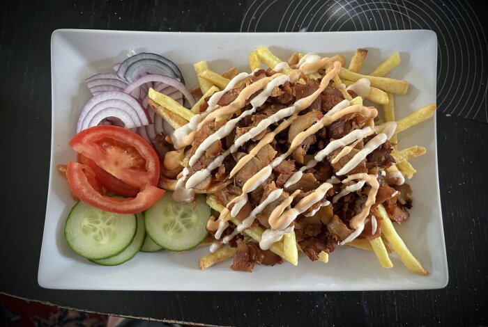 Tallrik med kycklingkebab, pommes och sås, toppad med tomat, rödlök och gurka.